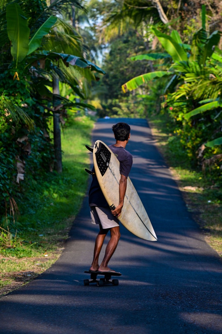 Surfer carrying his board while riding a surfskate down a village road in Medewi