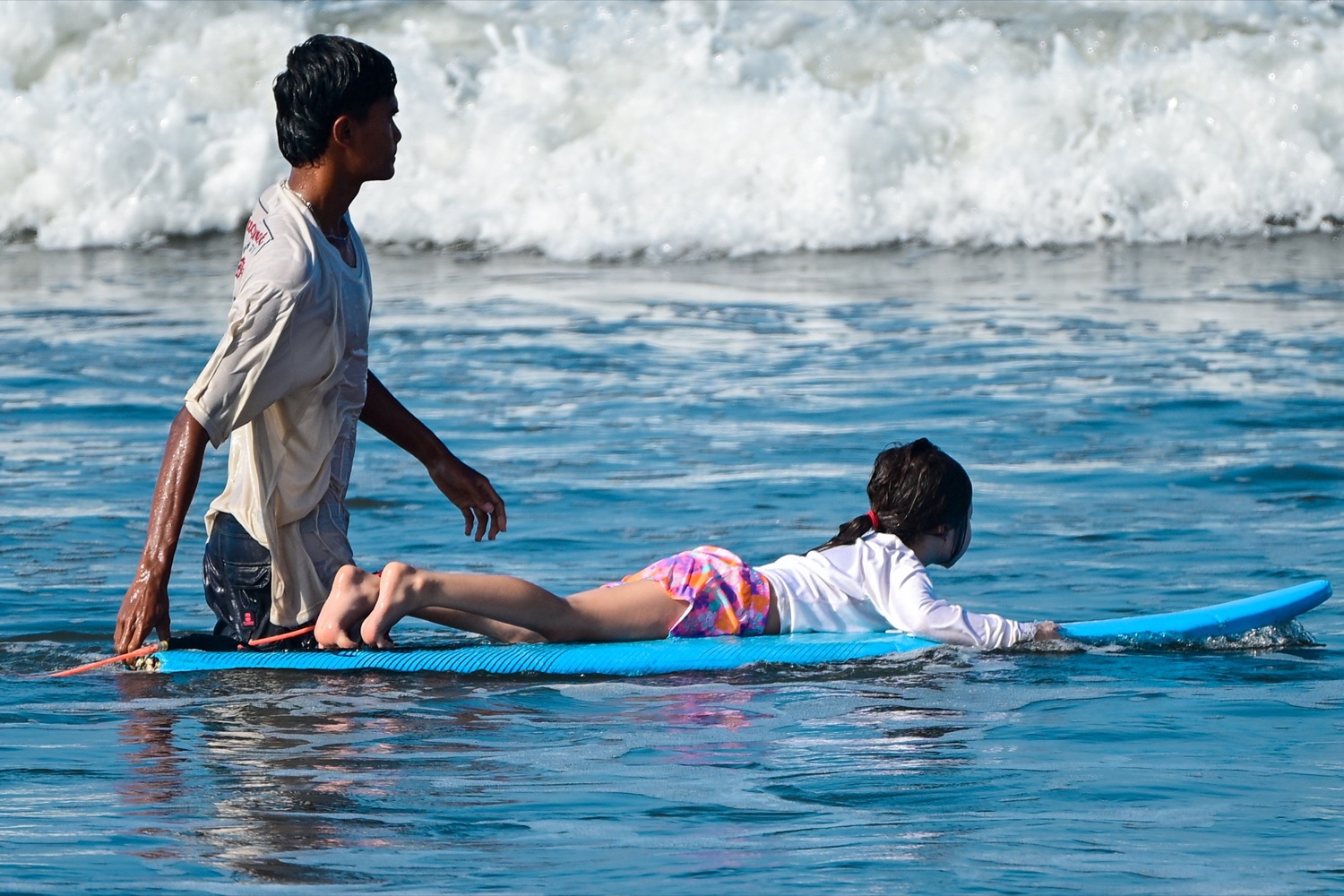 A coach standing in shallow water beside a small girl on a soft top at Medewi