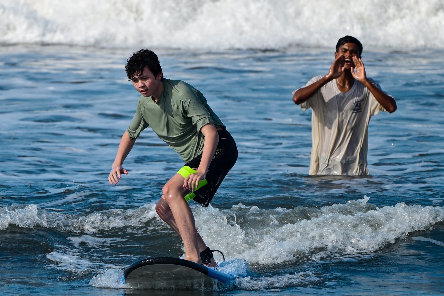 A young surfer riding a wave while his coach cheers from the water